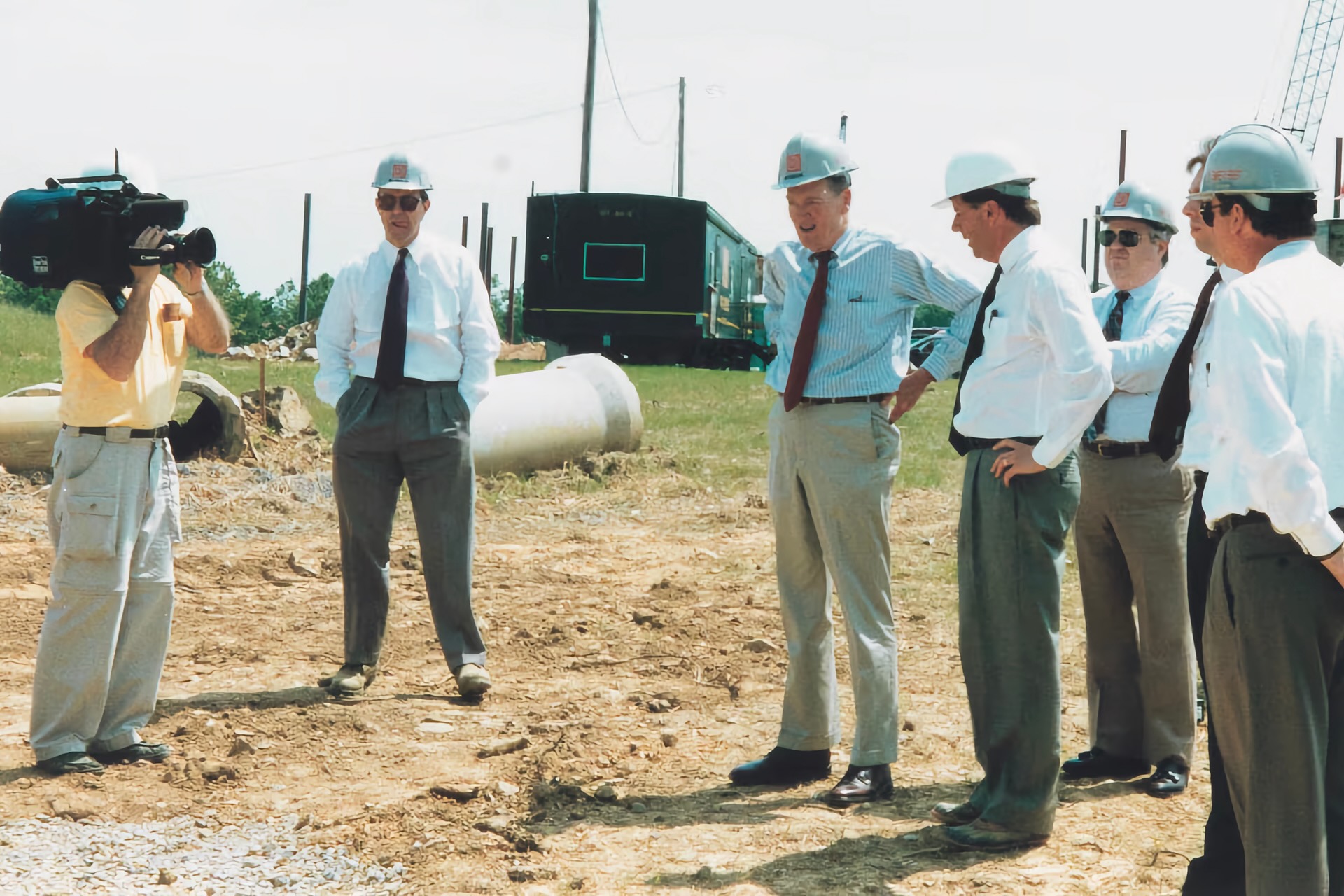 Historic photograph of six men in ties and hard hats inspecting a building site as a cameraman records the scene.