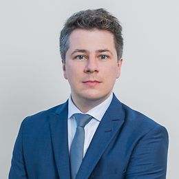 A man in a suit and tie smiles neutrally while looking into the camera 