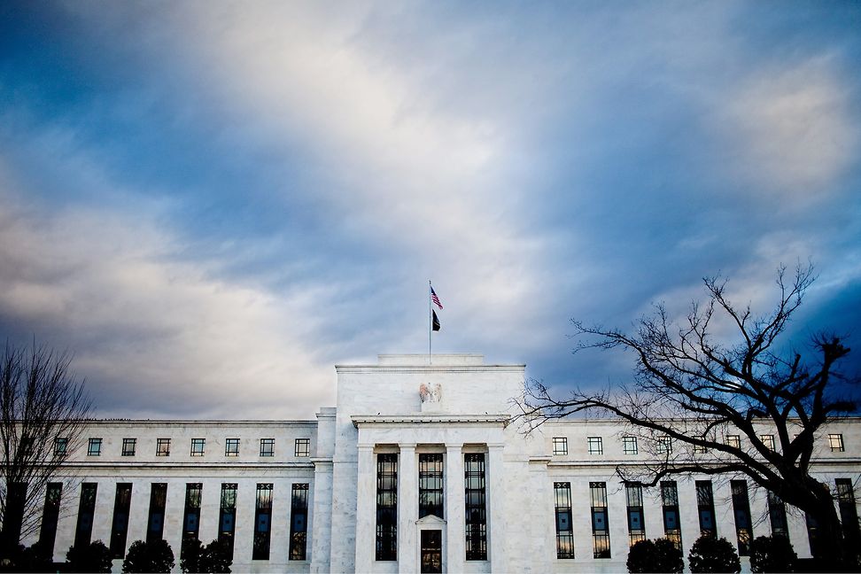 Outside view of a white building in striped classical architecture, crowned by an eagle. A US flag flies atop the building.