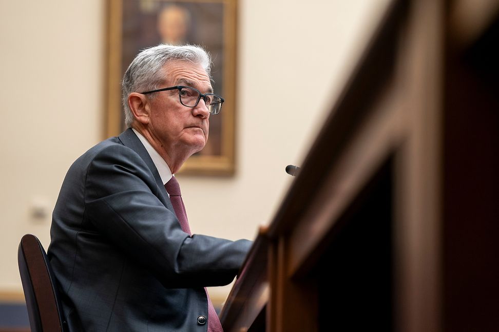 A grey-haired man sits at a large table in front of a microphone at a political hearing