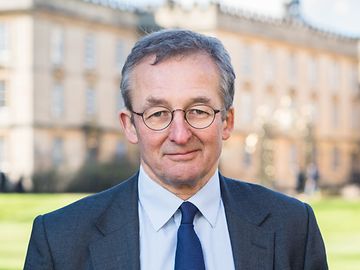 A man in a tie, jacket and glasses smiles friendly into the camera, a historic building in the background