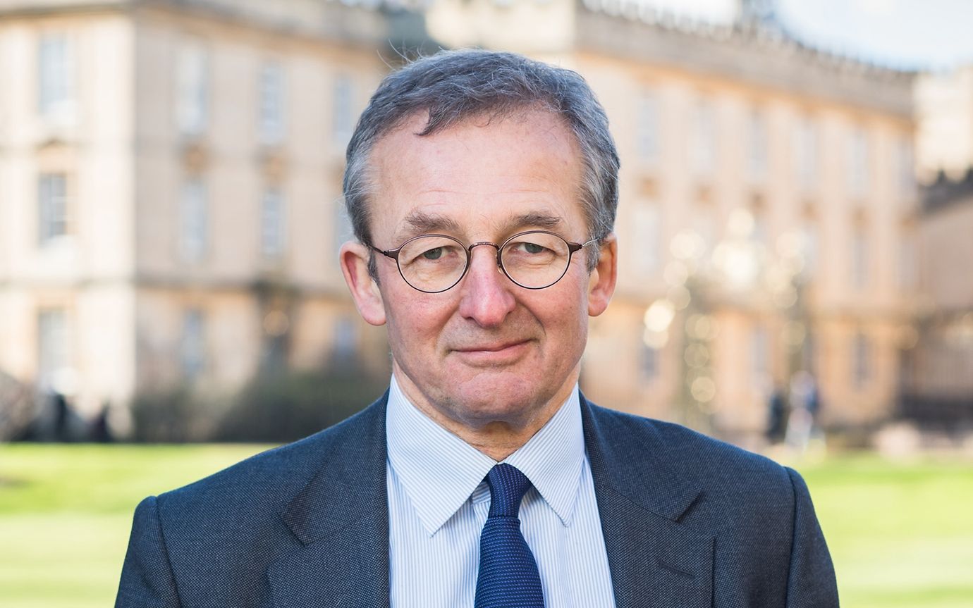 A man in a tie, jacket and glasses smiles friendly into the camera, a historic building in the background