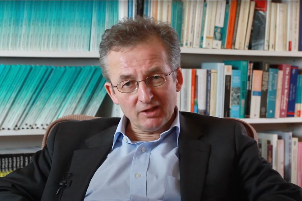 A man sits and talks, many books in the background