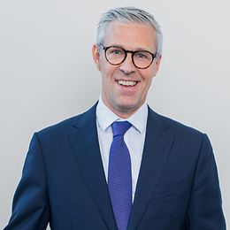 A man with glasses, jacket and tie smiles friendly into the camera