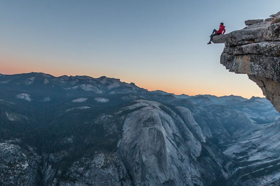 A person is sitting on a mountaintop, looking out over a mountain range that, due to perspective, appears to be at their feet.