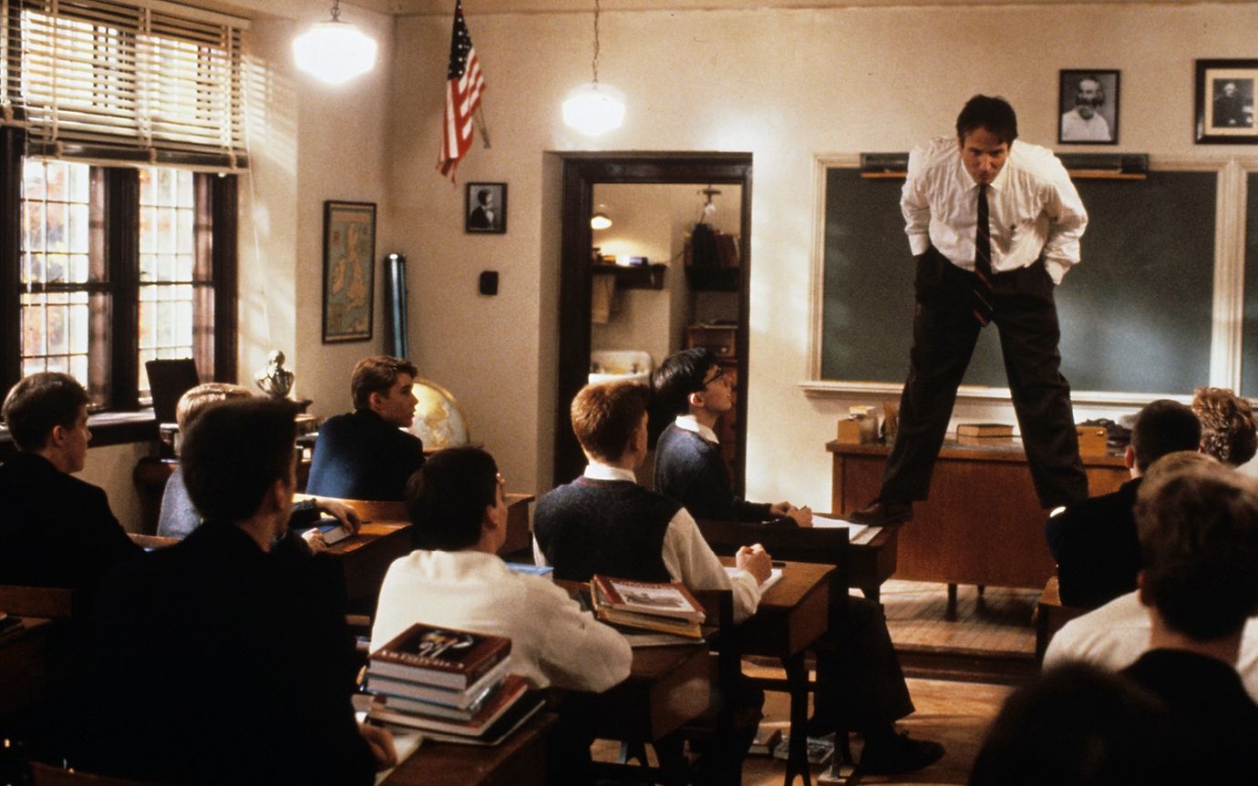 English teacher stands on top of a desk in a classroom, addressing students who are looking up at him.