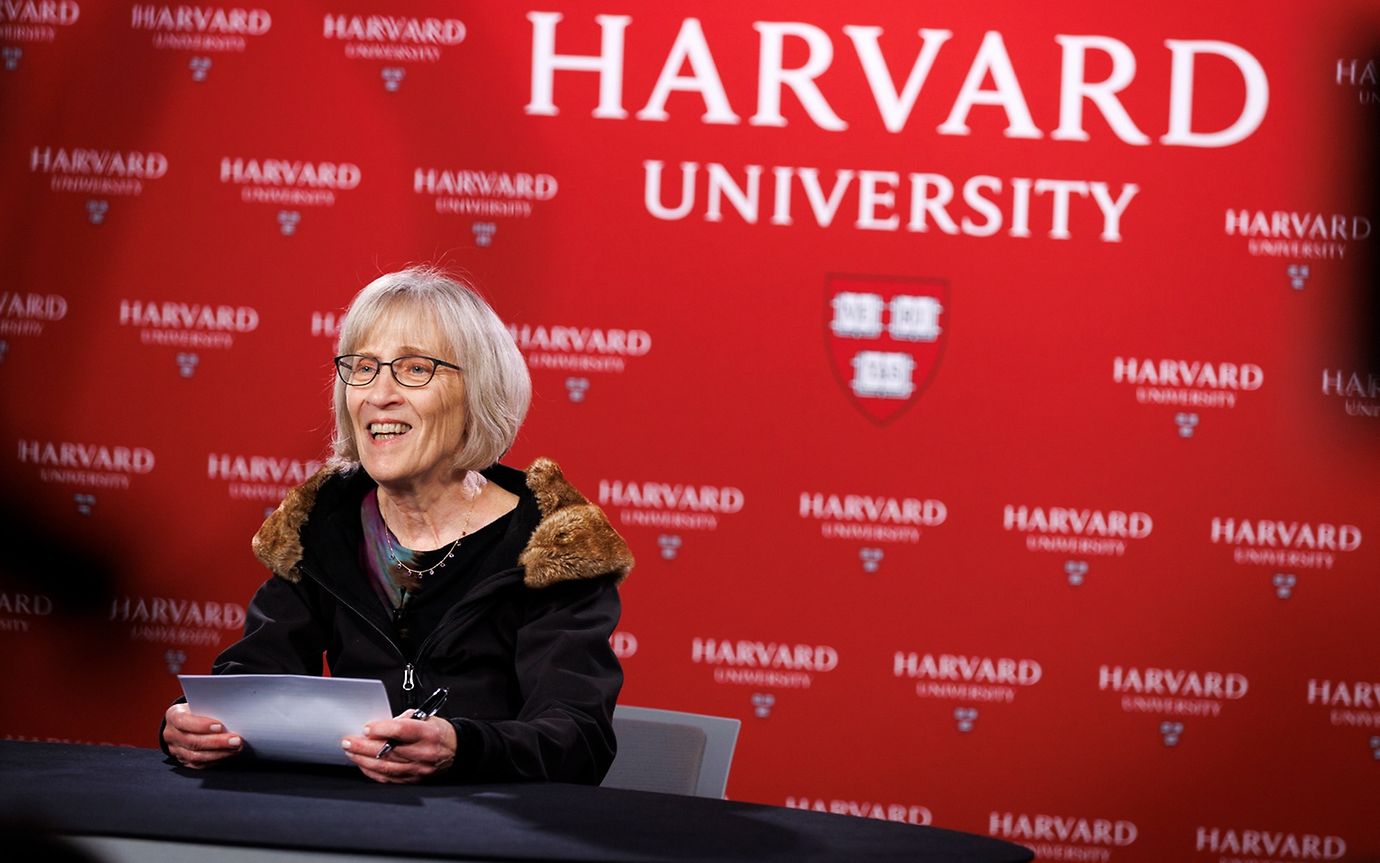 A woman with glasses and documents is speaking in front of a wall with the Harvard University logo.