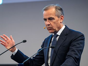 A man in a suit and tie speaks into a microphone and gestures with his hand.