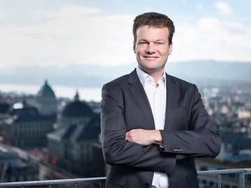 Reto Knutti, standing on a terrace with Lake Zurich and the university in the background, looks into the camera.