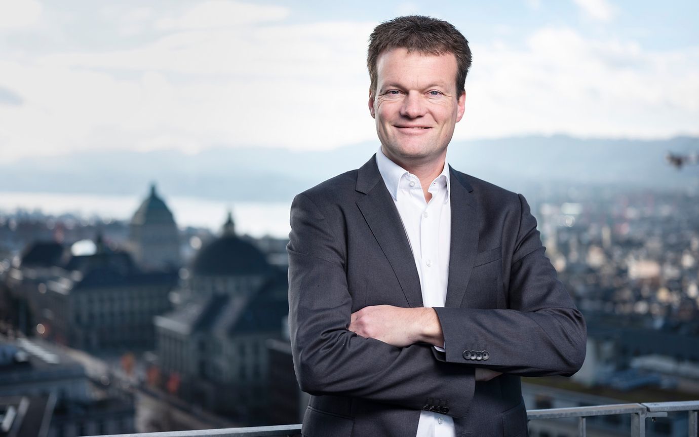 Reto Knutti, standing on a terrace with Lake Zurich and the university in the background, looks into the camera.