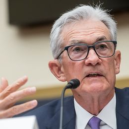 An elderly man wearing glasses, a suit and tie, speaks into a microphone and gestures with his hands.