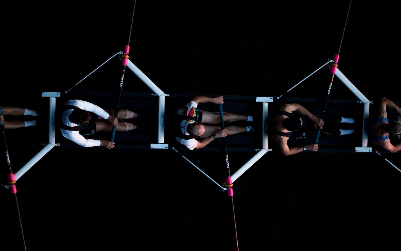 A bird's eye view of five athletic rowers in a skiff, standing out from the seemingly black surface of the water. 