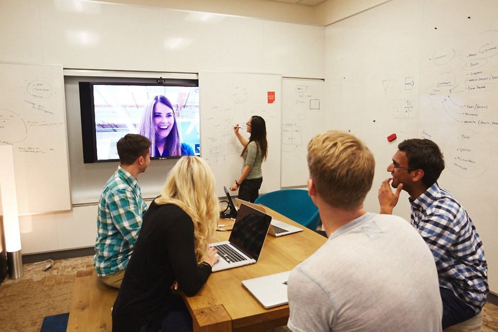 5 men and women are holding a virtual meeting sitting at a wooden table. A woman is connected via a large screen.