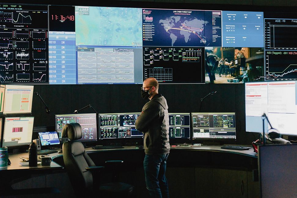 A man stands in front of a wall of screens displaying graphs, numbers and progressions.