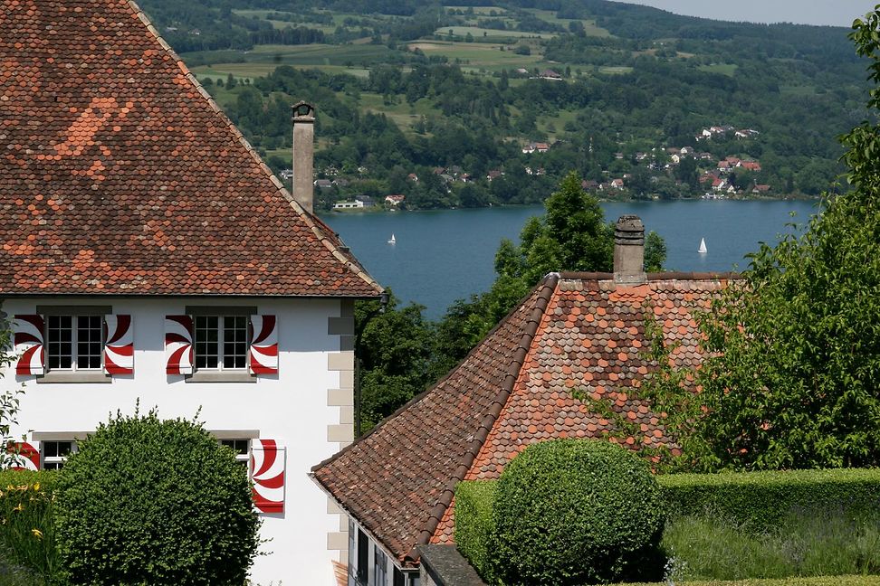 Cut to a castle-like building with red and white shutters, surrounded by spring-like trees.