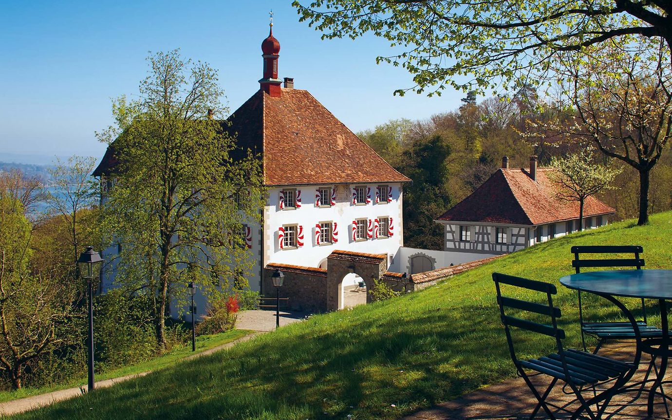 In spring, a small castle with red and white shutters nestles among trees and meadows above a lake. 