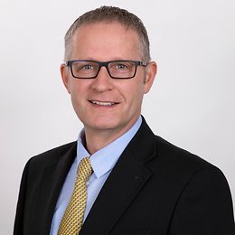 Middle-aged man with horn-rimmed glasses and wearing a dark suit with a yellow tie looks friendly into the camera.