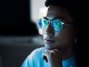 Close-up of a man, chin resting on his hand, with a computer screen reflected in his glasses.