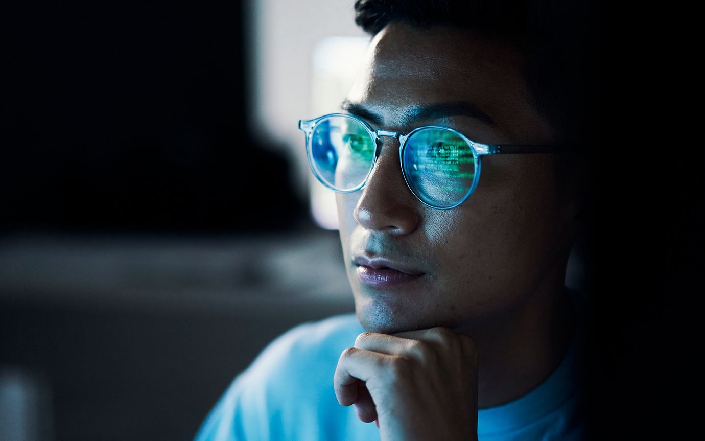 Close-up of a man, chin resting on his hand, with a computer screen reflected in his glasses.