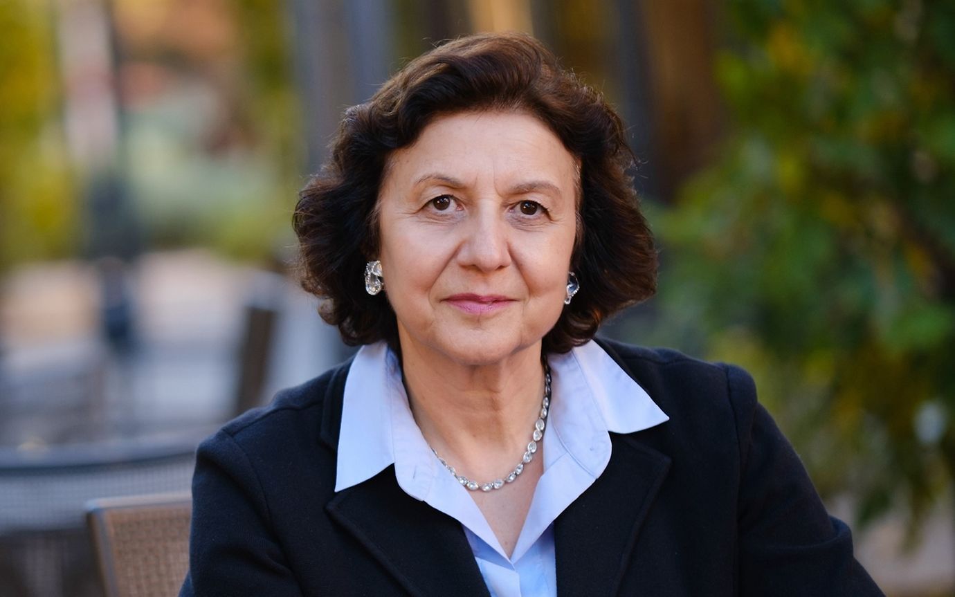 A woman in business attire is sitting at a table in a garden, her hands folded in front of her, looking into the camera.