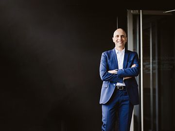 Middle-aged man in a suit, leaning against a wall with his arms crossed in front of a dark background.