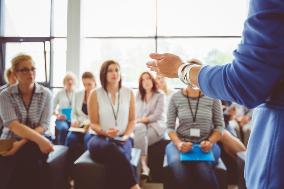 Cut from behind, you can see the left hand of a speaker addressing a mixed group of adults who are listening attentively.