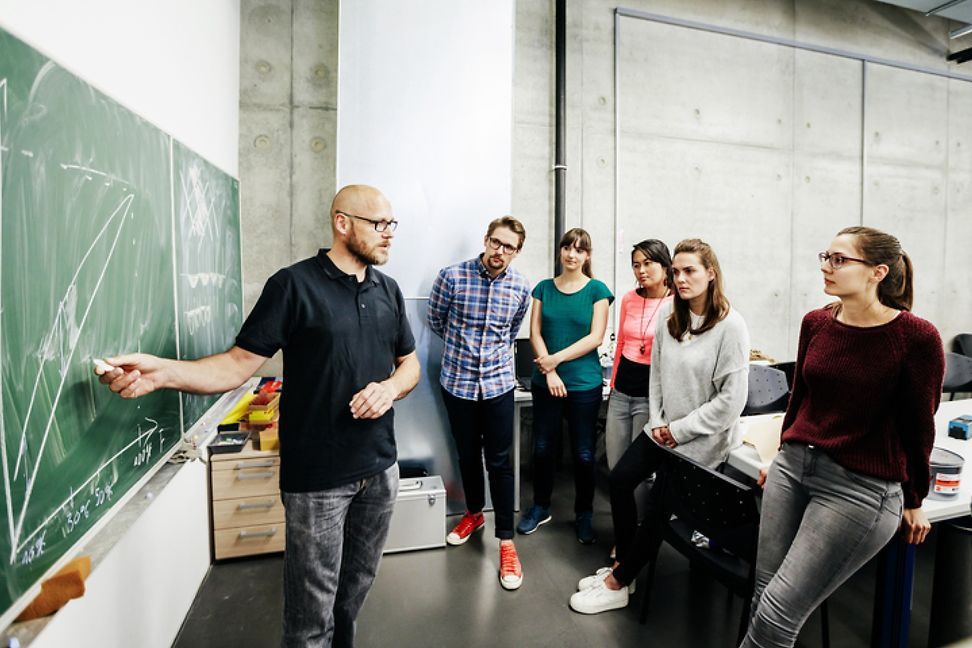 A man stands at a blackboard in a classroom, explaining a graphic to a diverse group.