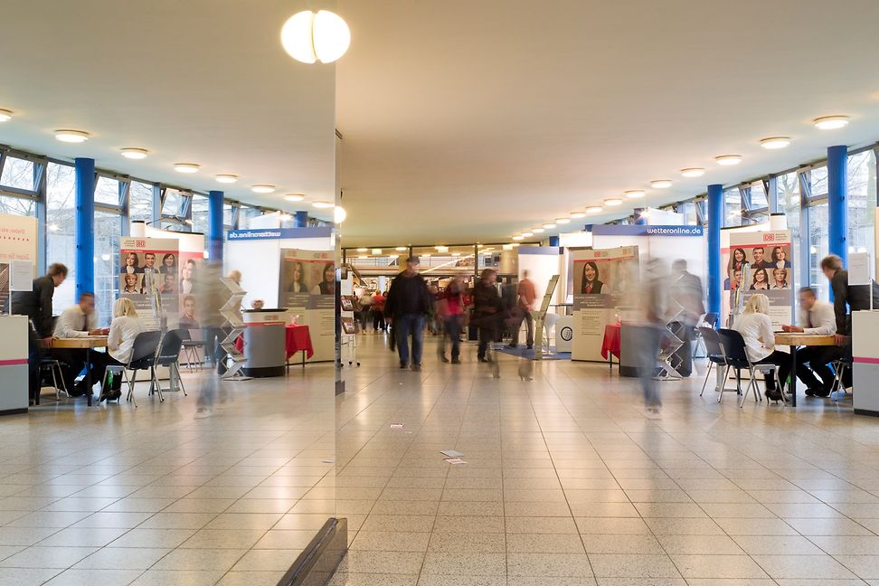 Smaller presentation stands in a large hall are surrounded by young people.