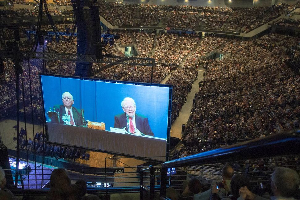 Thousands of people in a huge hall, while on a big screen two aged businessmen sit at a table and talk.