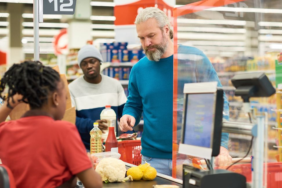 People stand in front of consumer goods on a conveyor belt while a person operates a screen and a scanner.