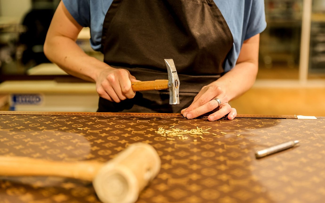 A person in work clothes in a workshop holds a hammer and ruler on a piece of leather marked 'LV'.