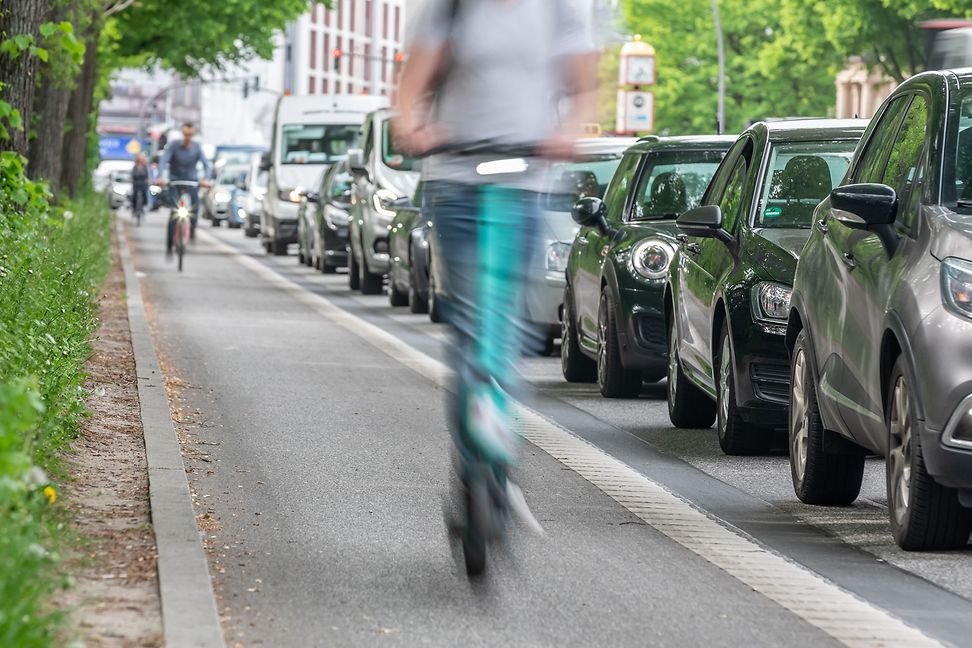 A person is riding a scooter on the pavement past a line of parked cars in a city.