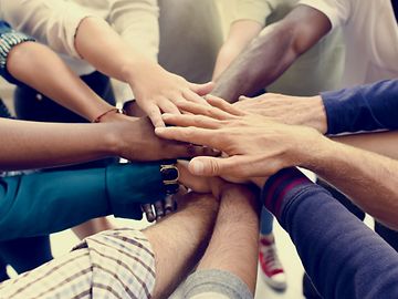 Many people of different colours stand in a circle and stretch out their arms so that their hands meet in the middle.