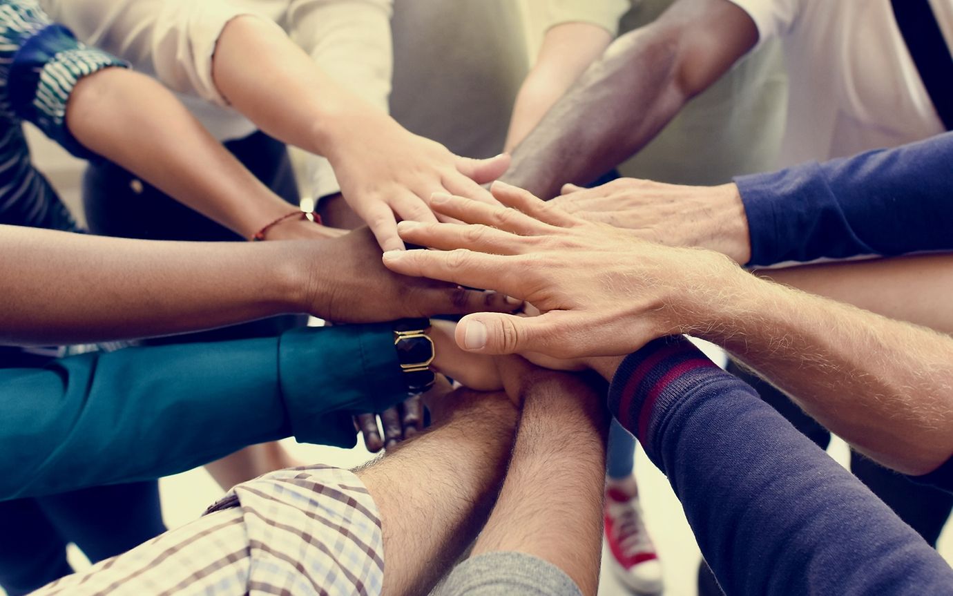 Many people of different colours stand in a circle and stretch out their arms so that their hands meet in the middle.