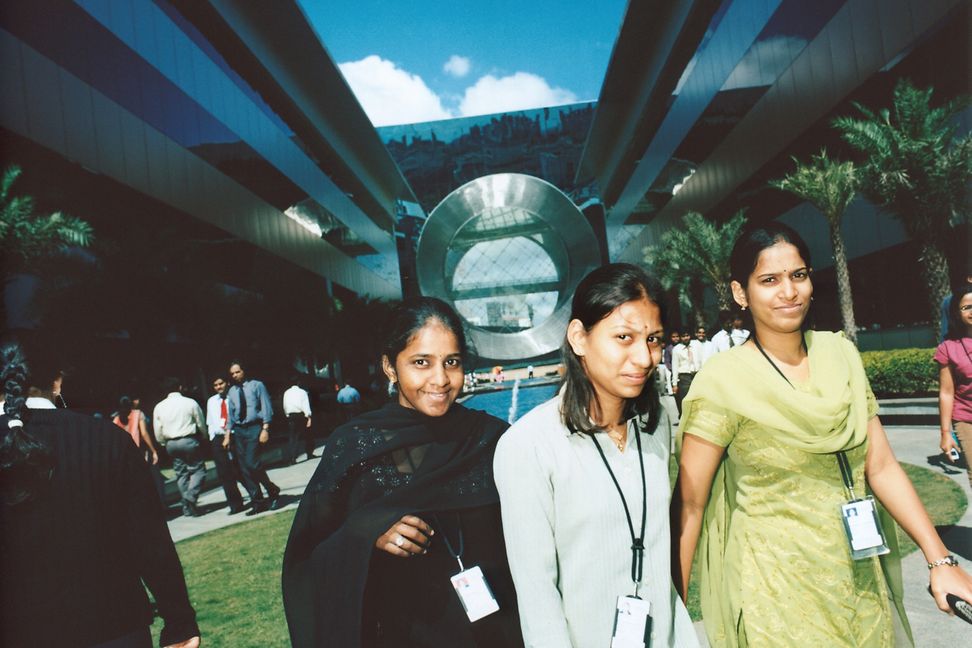 Three young women in saris and T-shirts walk in a leafy courtyard in front of large commercial buildings. 