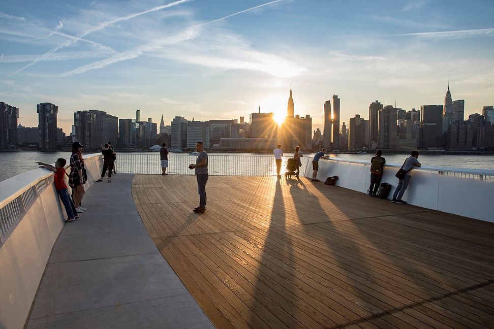 Five people stand against the evening sun on a wooden footbridge on the banks of a body of water, in the background a skyline. 