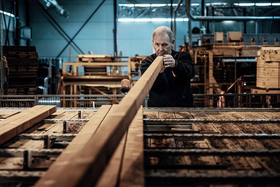 A man in a production hall holds a long beam in his hand and looks along the edge of the wood at the reader. 