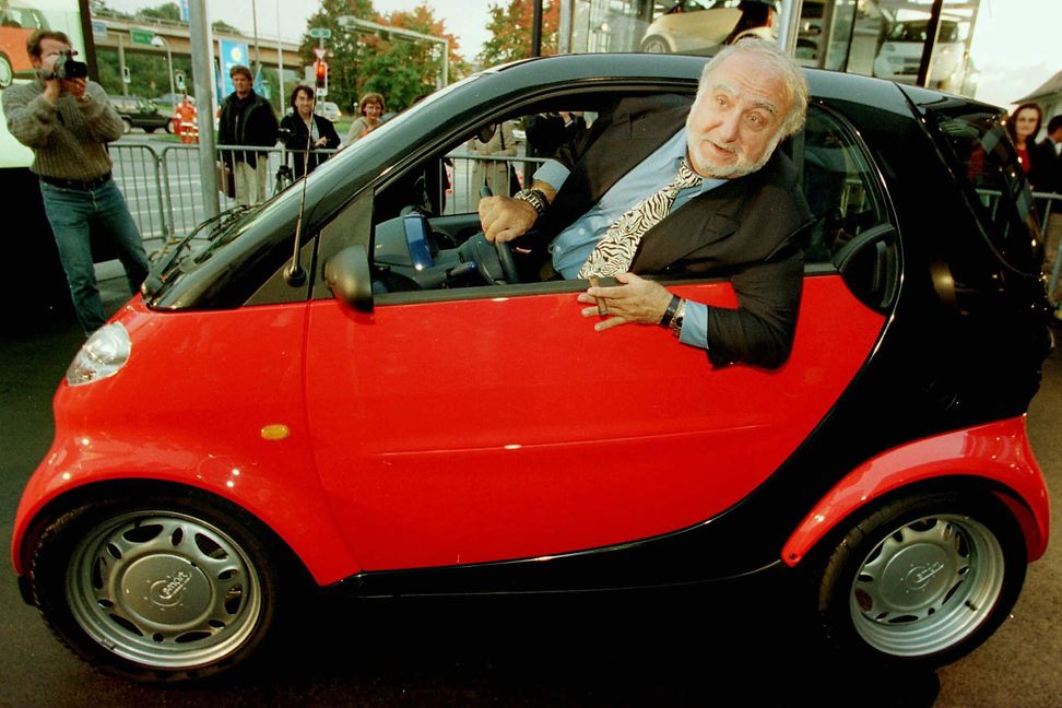 An elderly gentleman leans out of the window of a very small red car as people watch and take photographs.