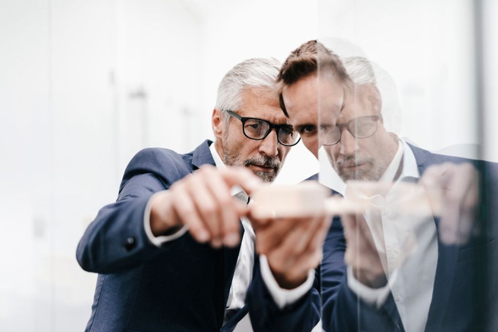 Two men in suits are holding and examining an object together, a gentleman is reflected in the glass.