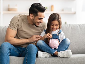 A man and a child are sitting on a sofa in a domestic setting, putting something into a pink clay pig.