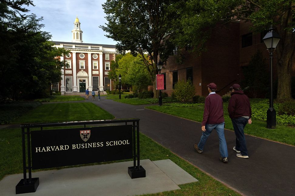 People are walking towards a brick building with a sign outside that says 'Harvard Business School'.