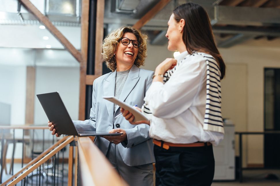 Two women, one with a laptop and the other with a notepad, talking in an office environment.