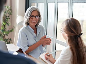 An older woman and a younger person with a notebook are exchanging ideas.