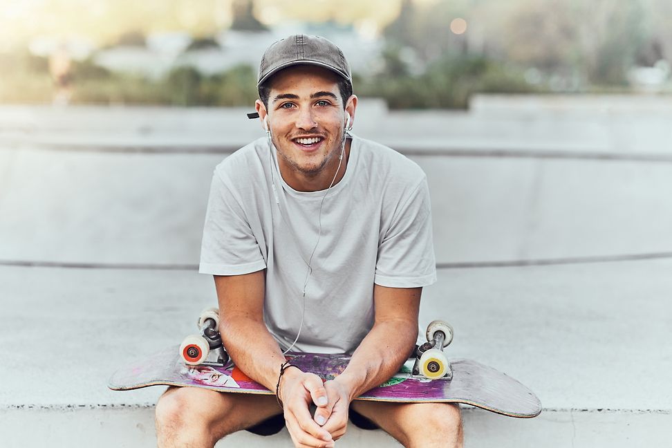 A young man sits with a skateboard on his lap and smiles at the camera.