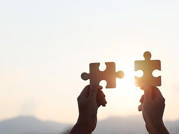 Hands are raised against a bright, white sky, each holding a piece of a wooden jigsaw puzzle