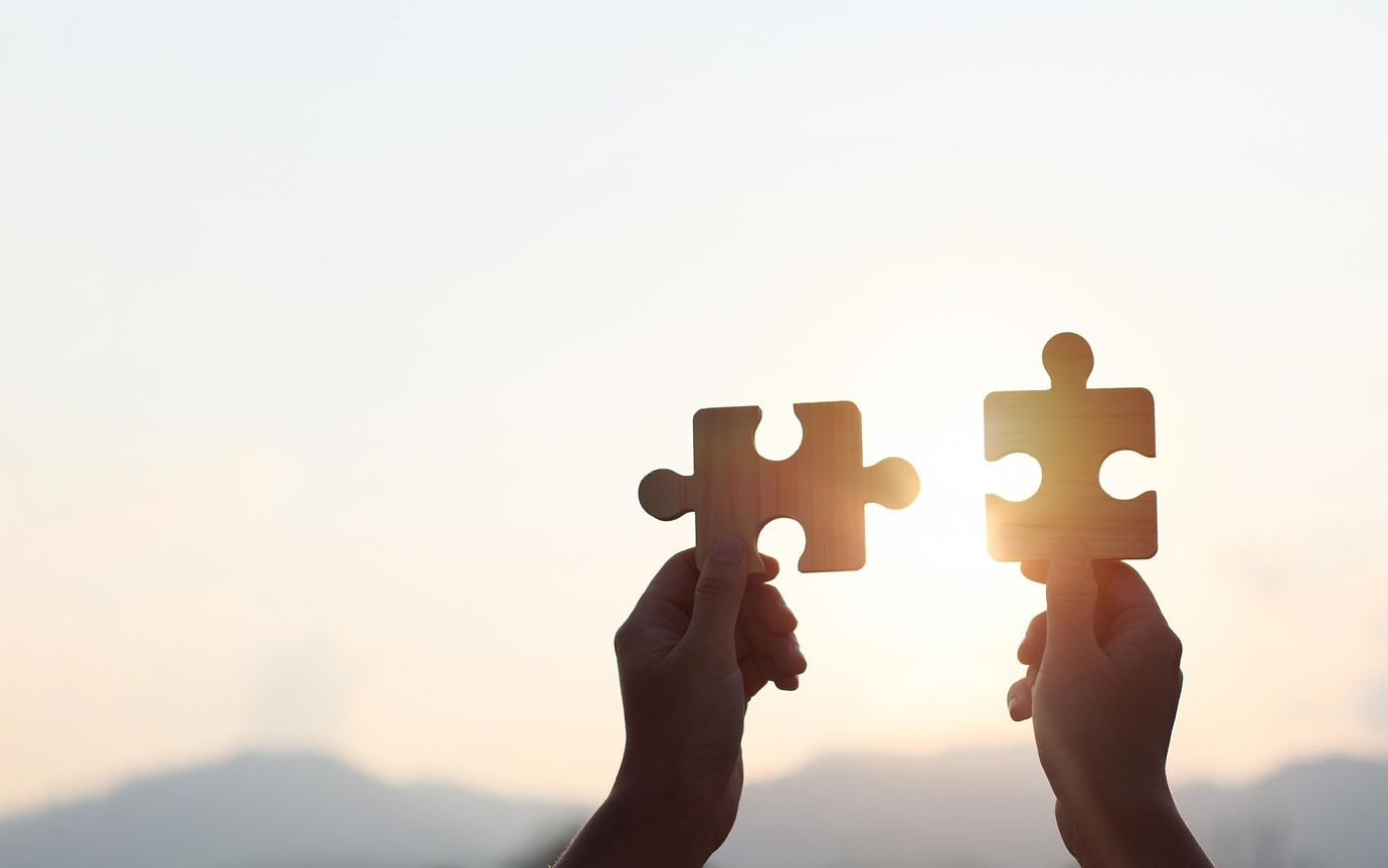 Hands are raised against a bright, white sky, each holding a piece of a wooden jigsaw puzzle