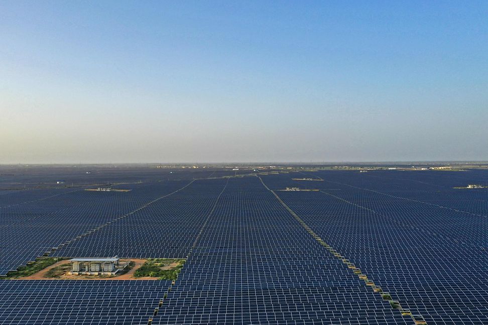 A large area of dark solar panels is interrupted on the left by a small building on a light brown background.